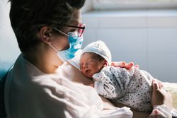 Mother wearing a face mask while holding her newborn baby in the hospital room.