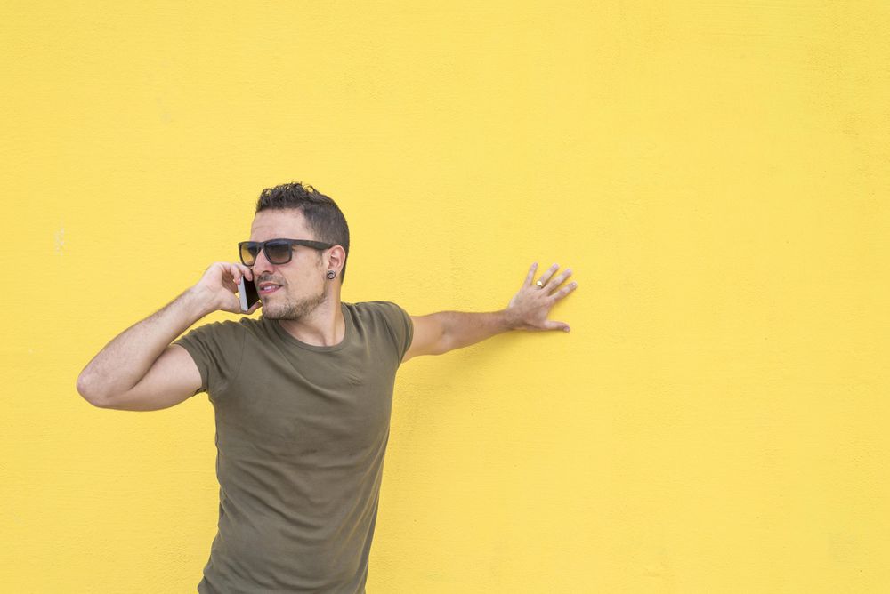 Serious male leaning on yellow wall talking on smartphone