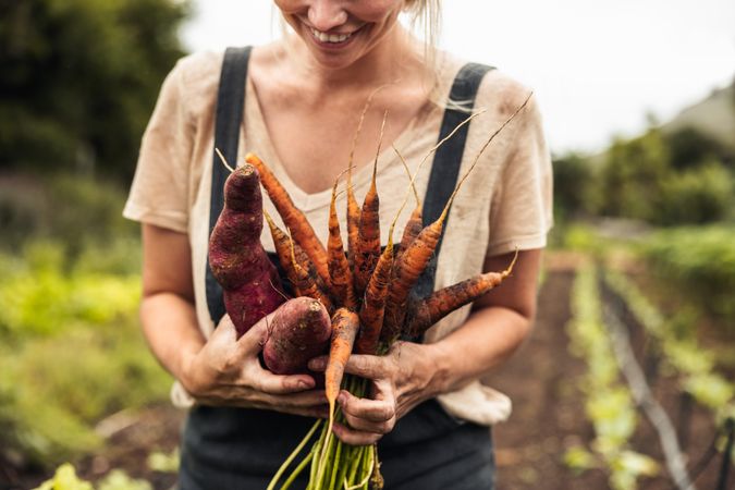 Happy female farmer holding freshly picked carrots and sweet potatoes on her farm
