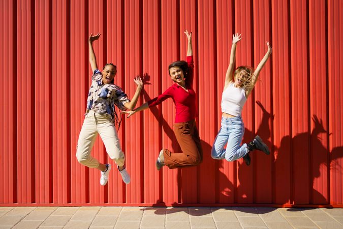 Three women jumping in front of red wall on sunny day