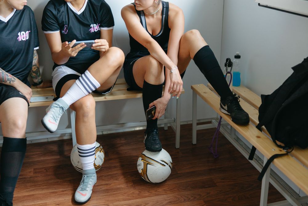 Group of teenage girls in soccer sports outfits sitting on wooden bench indoor