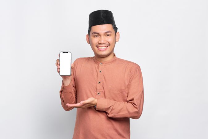 Happy Muslim man in kufi hat holding mobile phone and showing mock up screen