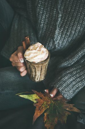 Woman in cozy sweater and jeans holding whipped cream topped drink with fall leaves