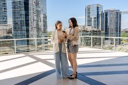 Two businesswomen standing on a terrace, one of them holding a cell phone and showing an email to her colleague. The scene is set in a city with tall buildings in the background.