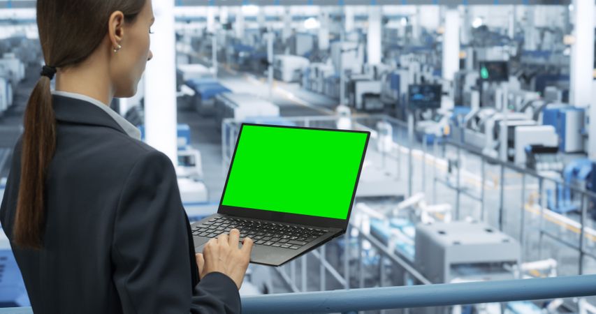 Young Female Engineer Using Laptop Computer with Green Screen Mock Up Display at an Electronics Factory. Automated Manufacturing Machines and AI Robots Are Monitored Online