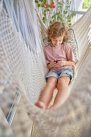 Adorable boy sitting in hammock with smartphone
