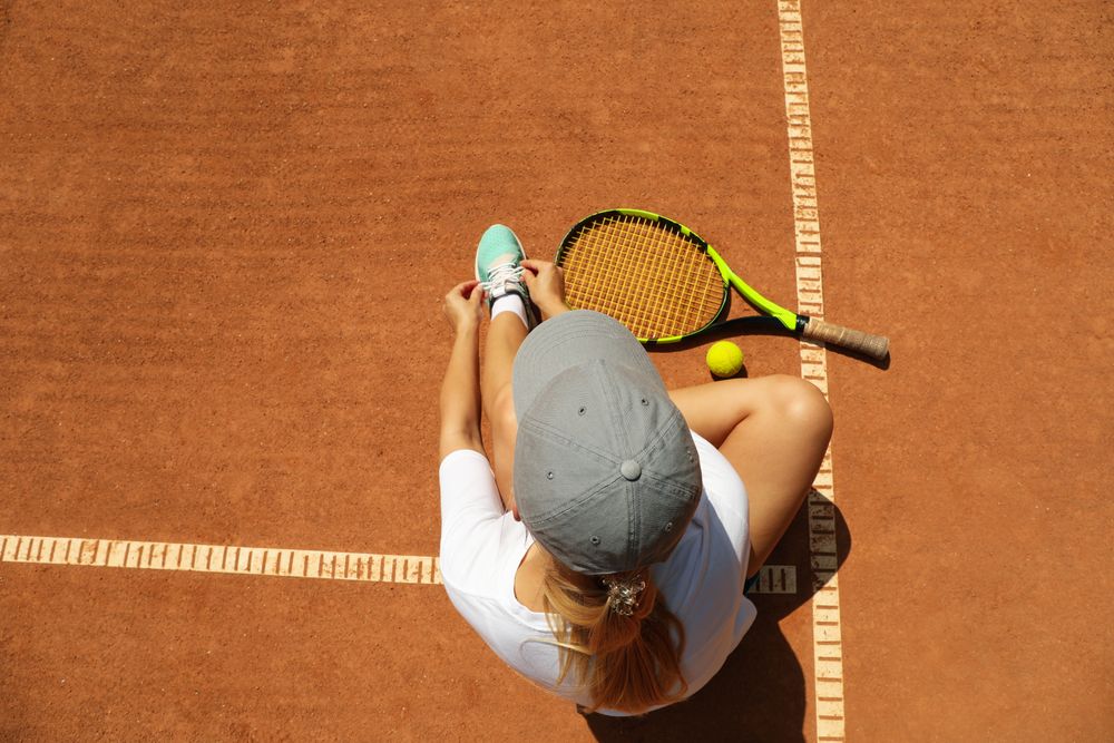 Woman ties shoelace on clay court with racket and and ball