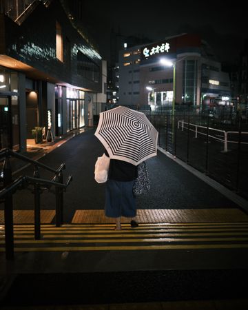 Back view of person holding an umbrella and standing on pedestrian lane during night time