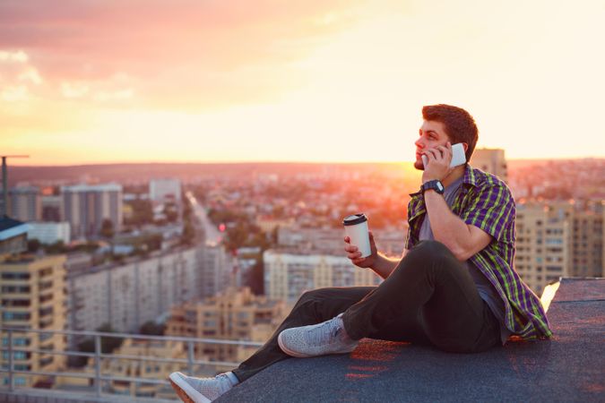 Male taking phone call while sitting on roof with cup of coffee