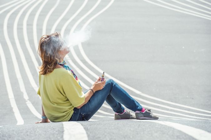 Modern relaxed woman vaping while sitting on marked road