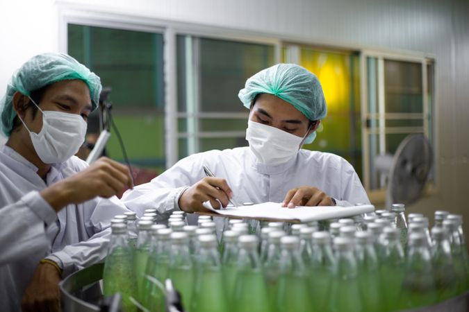 Male worker wearing protective uniform checking drink bottle on bottling machine following checklist in document.