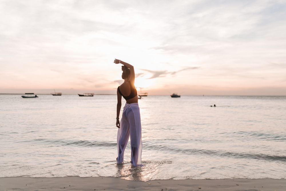 Back view of woman in dark bikini and light pants standing on beach at sunset