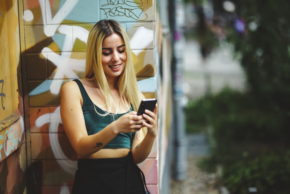 Stylish woman using smartphone at graffiti wall on city street.