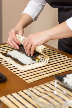 Hands of woman chef rolling up a Japanese sushi with rice, avocado and shrimps on nori seaweed sheet, vertical