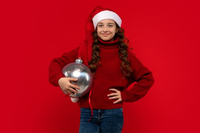 Girl in Santa hat and sweater poses with Christmas ornament.