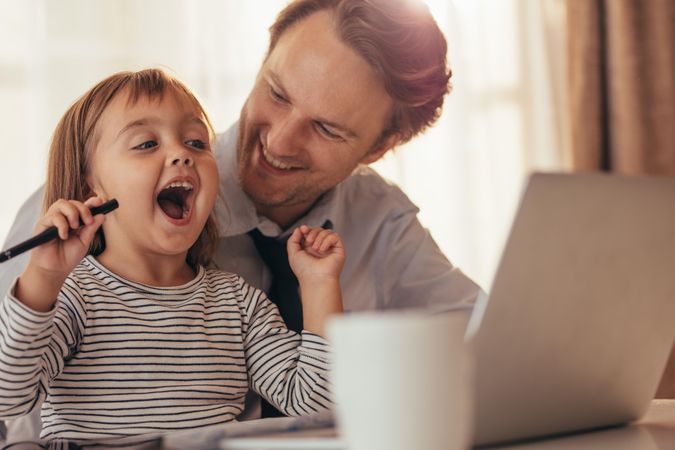 Girl playing with pen and laughing while sitting with her father at work with laptop