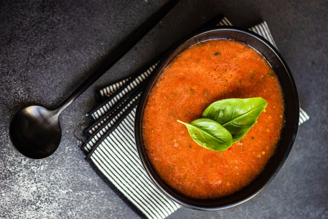 Top view of dark bowl of gazpacho soup with basil leaves on counter