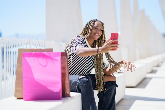 Happy female taking selfie seated outside in pedestrian area with shopping bags