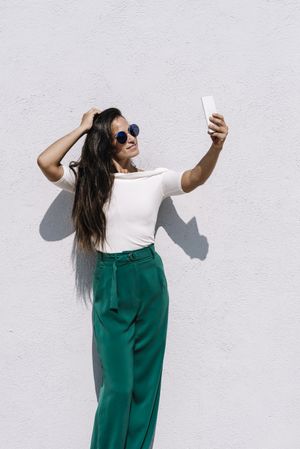 young female in trendy outfit posing for selfie while standing on bright background