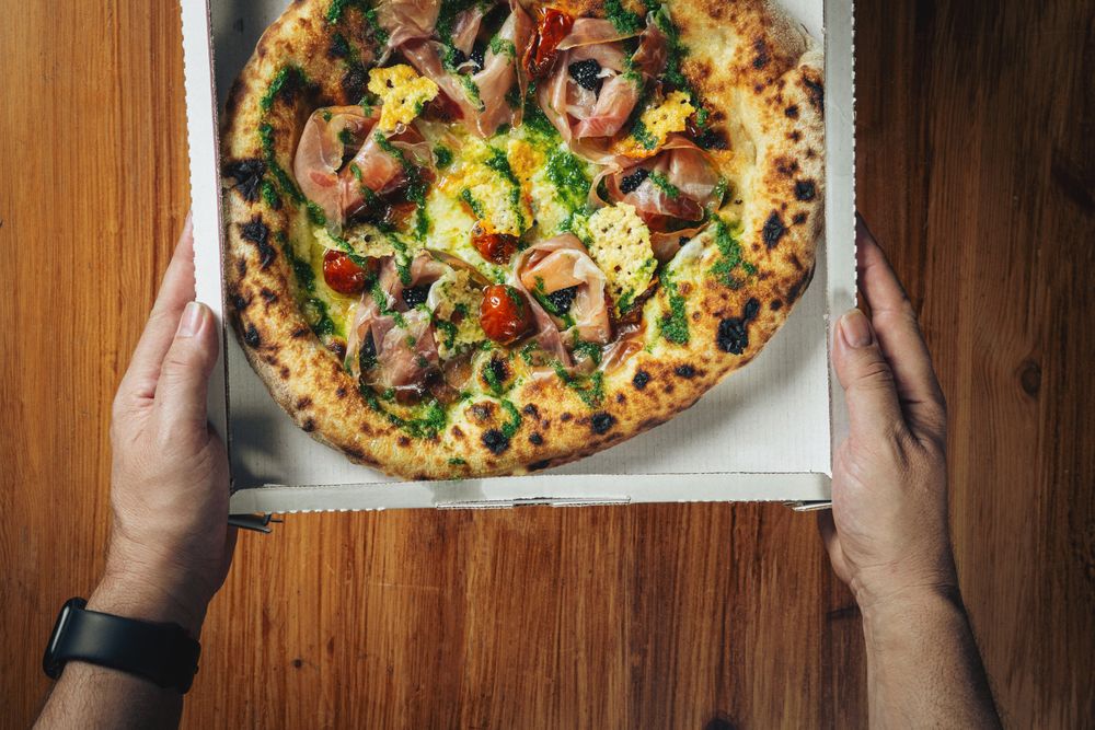Two gourmet pizzas with toppings in delivery boxes on wooden table surface