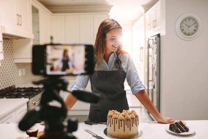 Smiling young woman standing at kitchen counter with pastries and camera