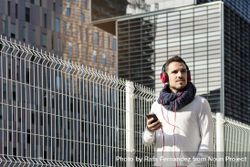 Young man walking in scarf past metallic fence and looking up from smartphone with copy space 4AzJ76