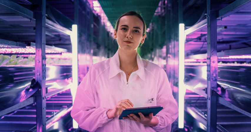 Portrait Female Farming Technician Standing Between Rows of Produce at a Modern Vertical Farm. Horticulturist Using Tablet Computer. Beautiful Young Woman Looking at Camera and Smiling