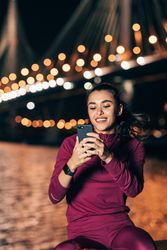 Woman in dark red sportswear smiling at phone near bridge at evening