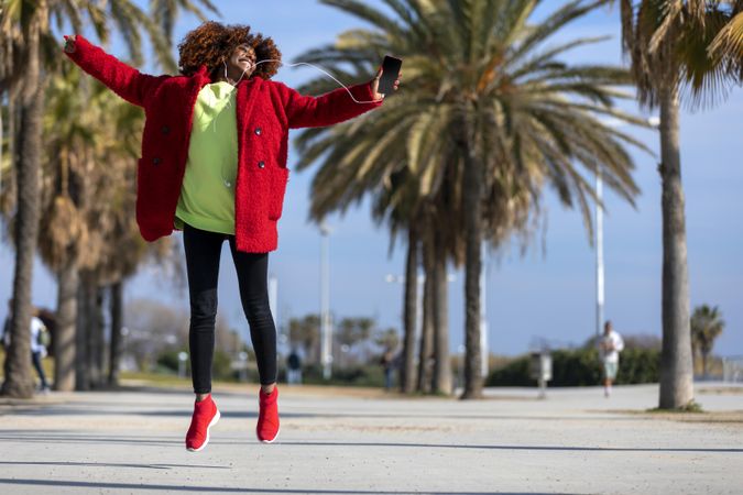 Female in bright colors jumping while listening to music outside on street with palm trees