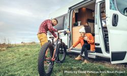 Female in orange sipping coffee sitting in camper van with male friend tending to bicycle bGygx5