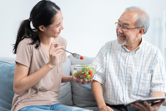 Female feeding mature male salad at home while he uses tablet