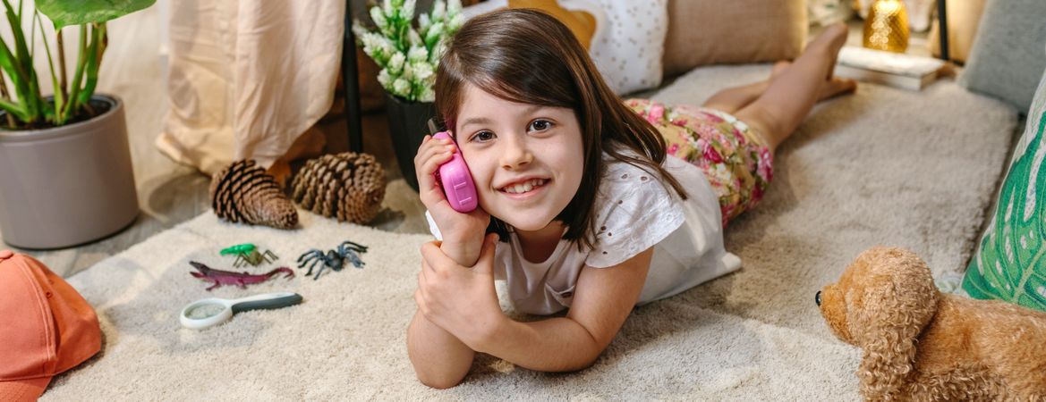 Banner of smiling little girl looking at camera while using walkie-talkie lying over rug at home