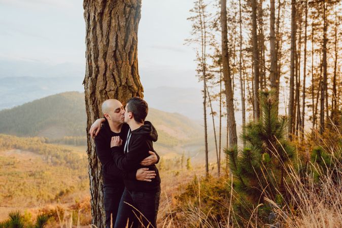 Smiling men hugging near tree in forest on hill