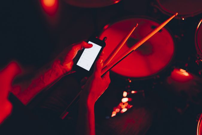 Man with drumsticks using smartphone near drums in redness