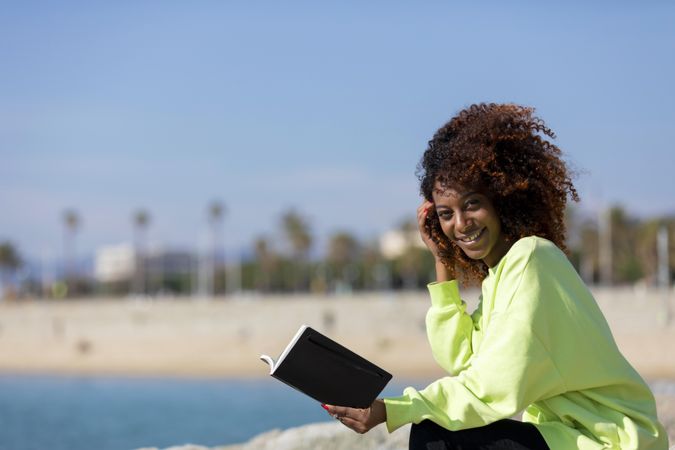 Smiling female in bright green shirt enjoying reading a book sitting by the ocean