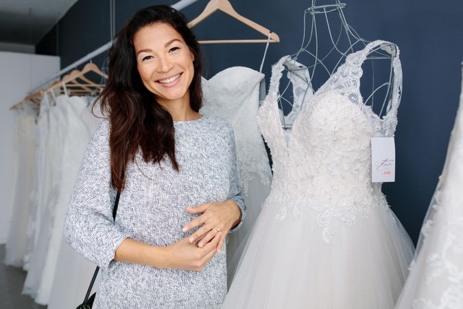Attractive young woman smiling and standing next to wedding dresses in bridal boutique