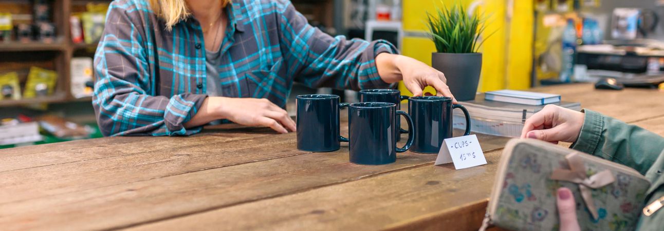 Banner of unrecognizable woman employee showing blue mugs to customer in vintage shop