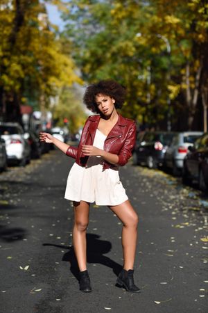 Serious woman in light one piece and red leather jacket standing in sunny road