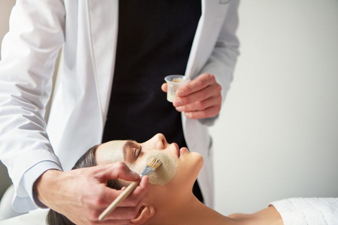 Woman having a mask painted on her face by man in lab coat