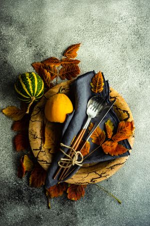 Top view of navy napkin, cutlery and gourds on wooden bowl and dried fall leaves