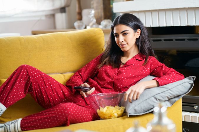 Female in red pajamas relaxing on sofa with bowl of chips and remote control