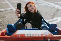 Smiling woman with smartphone in shopping trolley in parking lot
