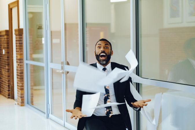 Laughing businessman in suit and tie celebrating by throwing papers in the air in office