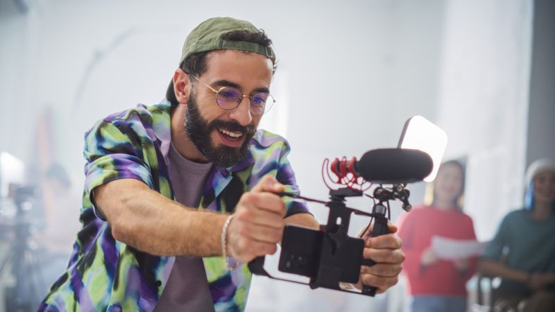 Young Male Filmmaker With Beard And Glasses, Joyfully Operating A Camera In A Busy Studio, Surrounded By Diverse Team Members Focused On A Creative Video Production Project.