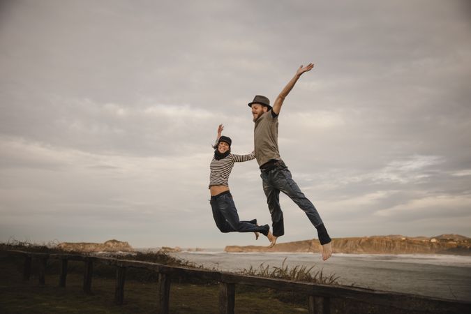 Couple holding hands and jumping on bench on shore near water
