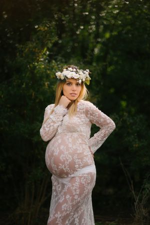 Pregnant woman with closed eyes in white lace dress touching chin while standing in forest