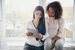 Two woman focused on a tablet together sitting by the window sill