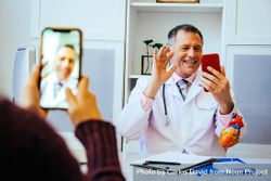 Smiling physician waving on video call with patient from his office 0vN770