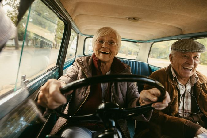 Happy older woman in warm clothing driving car with husband sitting in passenger seat
