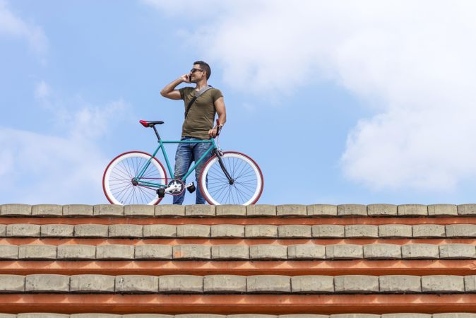 Male chatting on phone and standing with bike on roof with beautiful blue sky and clouds